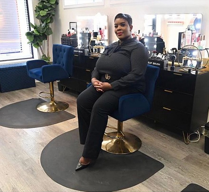 Jae Myers seated in a blue velvet chair in the salon, vanity mirrors and makeup behind her